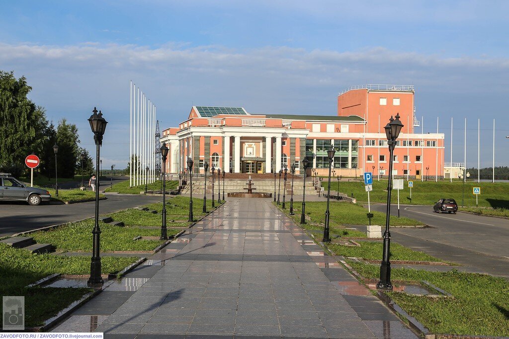 A wide, paved walkway leading toward a large, classical-style building with a red and white facade and columns at the entrance. The walkway is lined with lampposts and green grass. A car and a few people are visible in the distance.