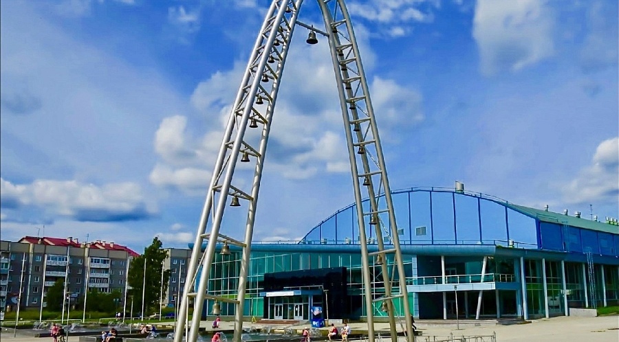 An outdoor public space with a modern, arch-like sculpture made of metal and featuring many bells suspended from it. In the background is a large, blue, arched building, and a few people are visible near a fountain in the foreground.