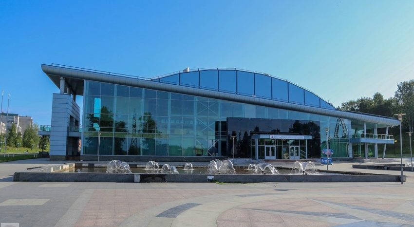 The exterior of a modern, multi-story building with a large glass facade. The building has a curved roofline and is surrounded by a paved plaza with a fountain. The sky is bright blue and clear.